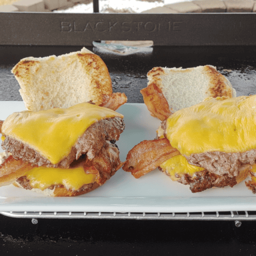 Close-up of two bacon smash burgers on a toasted bun, topped with melted cheddar cheese, served on a white plate with a Blackstone griddle in the background. A metal spatula is lifting one of the burgers, showcasing the crispy bacon and juicy smashed beef patties.