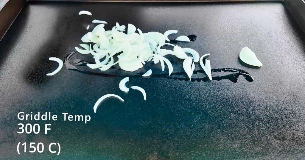 sliced white onions on a blackstone griddle with oil.  Showing the temperature of 300 degrees.