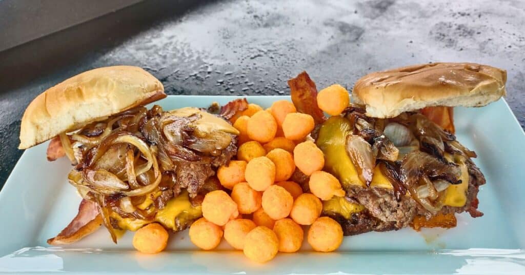 A plate of griddle-cooked cheeseburgers topped with caramelized onions and crispy bacon, served with a side of bright orange cheese puffs. The burgers are on toasted buns, slightly open-faced to showcase the melted cheese and toppings. The dish is presented on a light blue plate with a flat-top grill in the background.